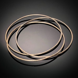 May include: Four gold-toned bangle bracelets stacked on top of each other. The bracelets are smooth and have a slightly hammered texture.