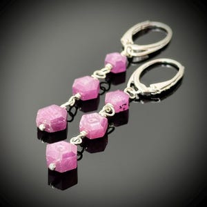 May include: Dangle earrings featuring faceted, pink gemstone beads strung on silver-toned wire. The earrings have a lever-back closure. The gemstones are a vibrant magenta color, and the earrings are displayed against a dark background.
