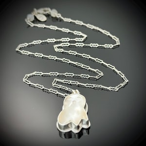 May include: A silver necklace with a ghost-shaped pendant. The pendant is white and has a silver border. The chain is made of small, interconnected links. The necklace is displayed on a black surface.