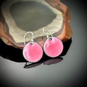 May include: A pair of pink enamel earrings with silver hooks. The earrings are round and have a black outline.