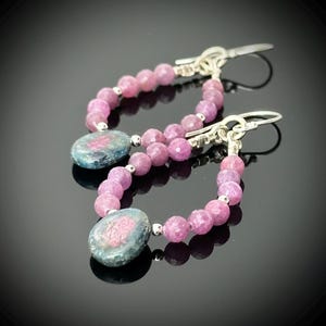 May include: A pair of dangle earrings featuring pink faceted beads and oval-shaped blue-green stones. The earrings have silver-colored hooks and small silver beads between the pink beads and the stones. The earrings are displayed on a black surface.