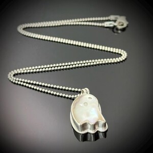 May include: A silver necklace with a ghost-shaped pendant. The pendant features a large, iridescent pearl set in a silver bezel. The chain is made of small, round silver beads. The necklace is displayed against a dark background.