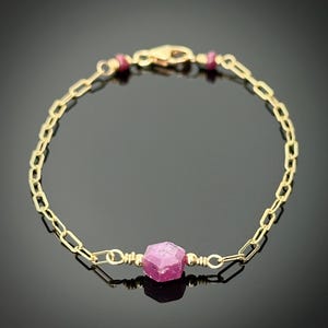 May include: A delicate gold-tone bracelet featuring a faceted, pink gemstone center. The bracelet has a chain link design with a lobster clasp closure. Small, round, pink gemstones accent the chain near the clasp.