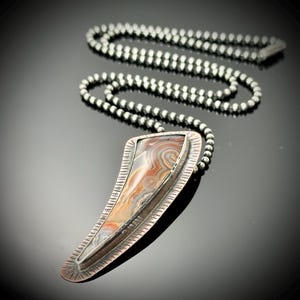 May include: A silver pendant necklace with a unique, elongated teardrop-shaped pendant. The pendant features a polished stone with swirling patterns in shades of brown, cream, and gray. The necklace chain is made of small, round silver beads.