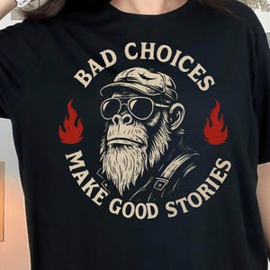 Puede incluir: Camiseta negra con un gráfico de un gorila con gafas de sol y gorra, con el texto "BAD CHOICES MAKE GOOD STORIES" en un diseño circular. Llamas rojas flanquean la imagen del gorila. Una prenda casual.