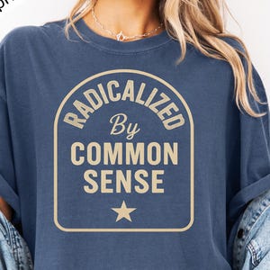 May include: A navy blue t-shirt with a tan graphic that reads "Radicalized by Common Sense" in a vintage style font. The graphic is surrounded by a tan border and features a tan star.