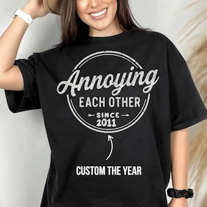 Könnte beinhalten: Ein schwarzes kurzärmeliges T-Shirt mit einer weißen kreisförmigen Grafik mit dem Text Annoying EACH OTHER SINCE 2011 und einem Pfeil, der auf den Text CUSTOM THE YEAR zeigt. Das Design hat eine abgenutzte Textur und ist vorne aufgedruckt.