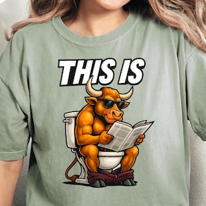 May include: Sage green t-shirt featuring the text "THIS IS" above a cartoon bull wearing sunglasses and reading a newspaper while on the toilet. The bull is brown with white horns.