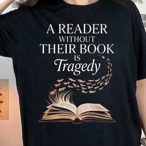May include: Black t-shirt with the text "A READER WITHOUT THEIR BOOK IS Tragedy" in white and gold. The design features an open book with a stream of stylized birds. The shirt is a casual wear item.
