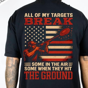 May include: Black t-shirt with a red and white graphic of a shooter aiming at a clay target, set against an American flag design. Text reads "ALL OF MY TARGETS BREAK" and "SOME IN THE AIR SOME WHEN THEY HIT THE GROUND."