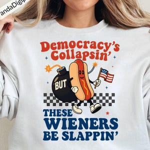 May include: White sweatshirt with a graphic featuring a cartoon hot dog holding an American flag, a bomb, and the text "Democracy's Collapsin' BUT THESE WIENERS BE SLAPPIN'". The design includes stars and a checkered pattern.