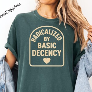 May include: Dark teal t-shirt with the text "RADICALIZED BY BASIC DECENCY" in a beige rectangular design with a heart. The brand name "PandaDigiprints" is in the upper left corner. The shirt has rolled-up sleeves.