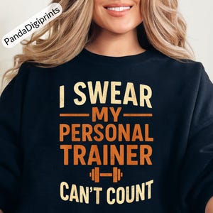 Puede incluir: Una sudadera negra con un gráfico de estilo vintage que dice "Juro que mi entrenador personal no puede contar" con un icono de mancuerna.