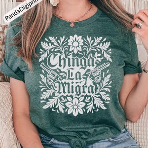 May include: Green t-shirt featuring a white floral design and the text "Chinga La Migra". The design includes two revolvers. The shirt is made of a soft material.