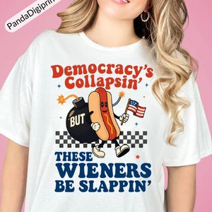 May include: White t-shirt featuring a cartoon hot dog holding an American flag, with the text "Democracy's Collapsin' BUT These Wieners Be Slappin'". The design includes a bomb and a checkered pattern.