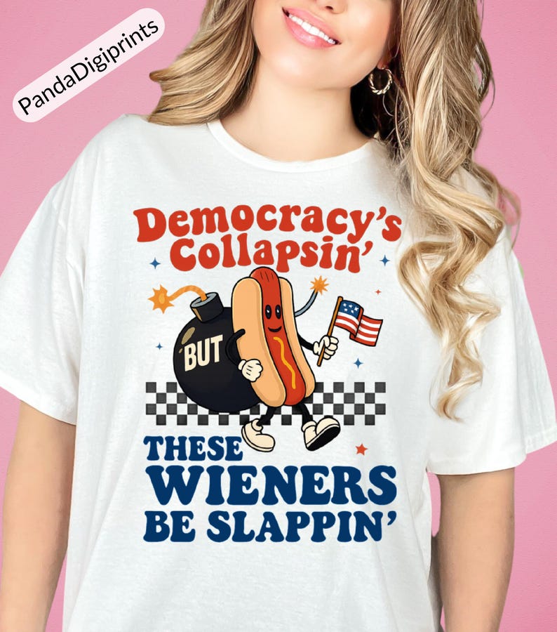 May include: White t-shirt featuring a cartoon hot dog holding an American flag, with the text "Democracy's Collapsin' BUT These Wieners Be Slappin'". The design includes a bomb and a checkered pattern.