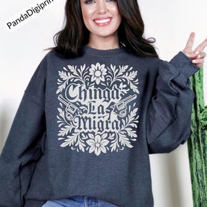 May include: Dark grey sweatshirt featuring a white floral and gun design. The text "Chinga La Migra" is displayed in a decorative font. The sweatshirt has a crew neck and long sleeves.
