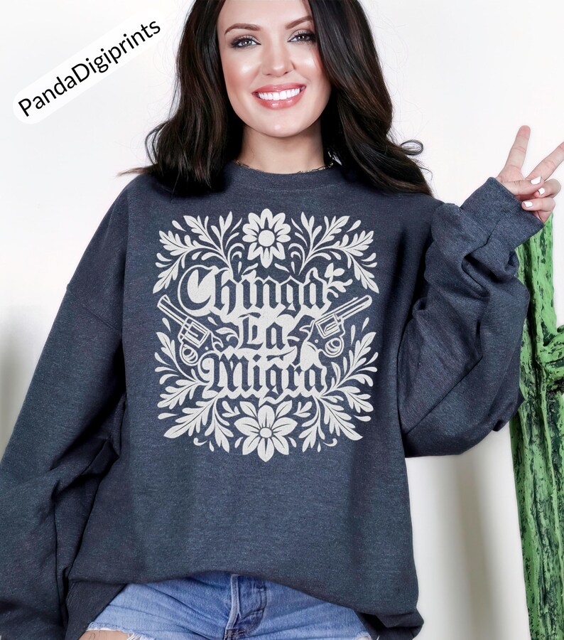 May include: Dark grey sweatshirt featuring a white floral and gun design. The text "Chinga La Migra" is displayed in a decorative font. The sweatshirt has a crew neck and long sleeves.