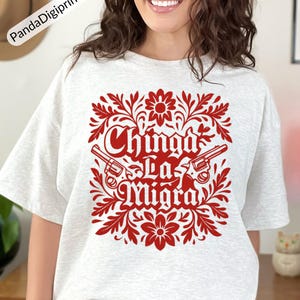 May include: A light grey t-shirt with a red floral design and the text "Chinga La Migra" and two revolvers. The design is centered on the shirt and is surrounded by floral elements.