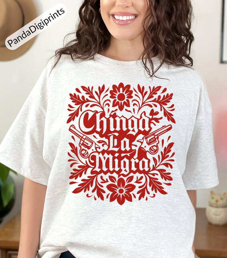 May include: A light grey t-shirt with a red floral design and the text "Chinga La Migra" and two revolvers. The design is centered on the shirt and is surrounded by floral elements.