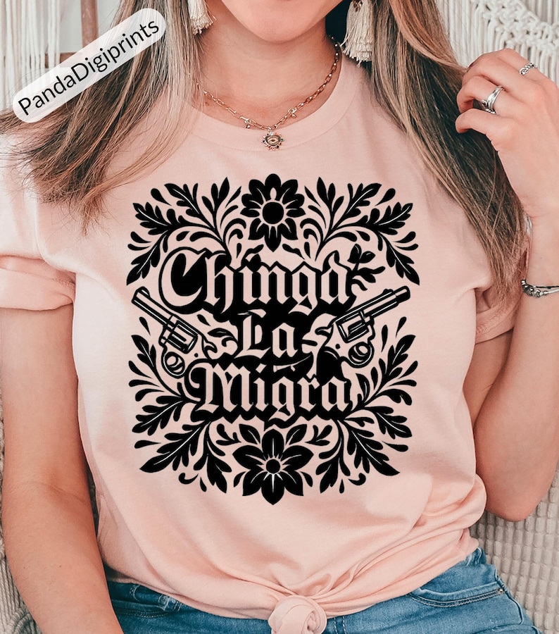 May include: A pale pink t-shirt with a black floral design and the words "Chinga La Migra" and two revolvers. The design is centered. The t-shirt is a crew neck style.