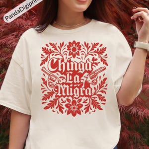 May include: A cream-coloured t-shirt with a red graphic. The design includes the text "Chinga La Migra", two revolvers, and floral motifs. The shirt is a casual, short-sleeved style, suitable for everyday wear.