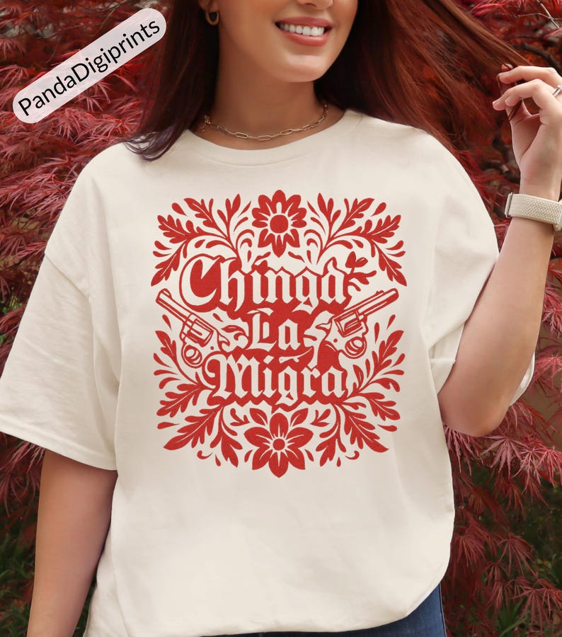 May include: A cream-coloured t-shirt with a red graphic. The design includes the text "Chinga La Migra", two revolvers, and floral motifs. The shirt is a casual, short-sleeved style, suitable for everyday wear.