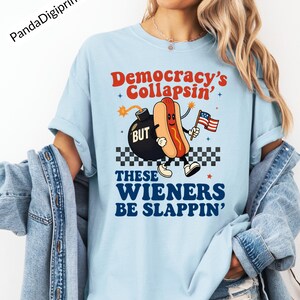 May include: Light blue t-shirt with a graphic of a hot dog holding an American flag, with the text "Democracy's Collapsin' BUT These Wieners Be Slappin'". The design includes a bomb and a checkered pattern.