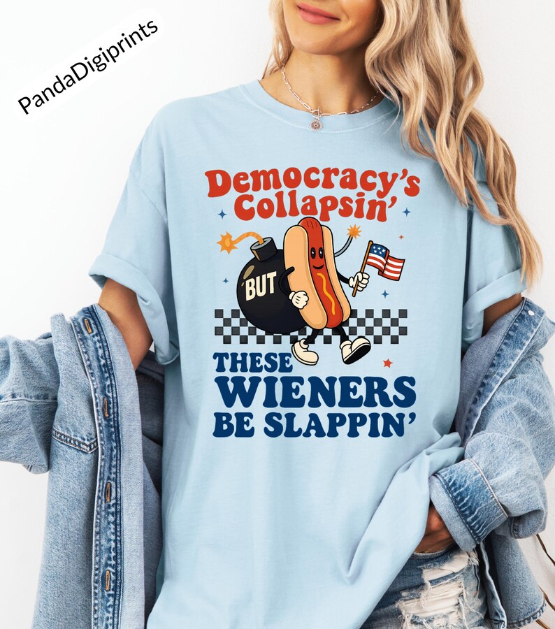 May include: Light blue t-shirt with a graphic of a hot dog holding an American flag, with the text "Democracy's Collapsin' BUT These Wieners Be Slappin'". The design includes a bomb and a checkered pattern.