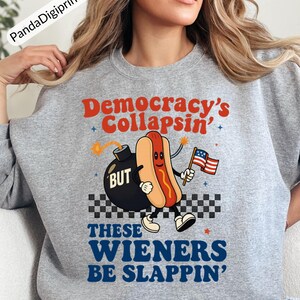 May include: A gray sweatshirt with the text "Democracy's Collapsin' BUT THESE WIENERS BE SLAPPIN'". The graphic features a cartoon hot dog holding an American flag next to a bomb.