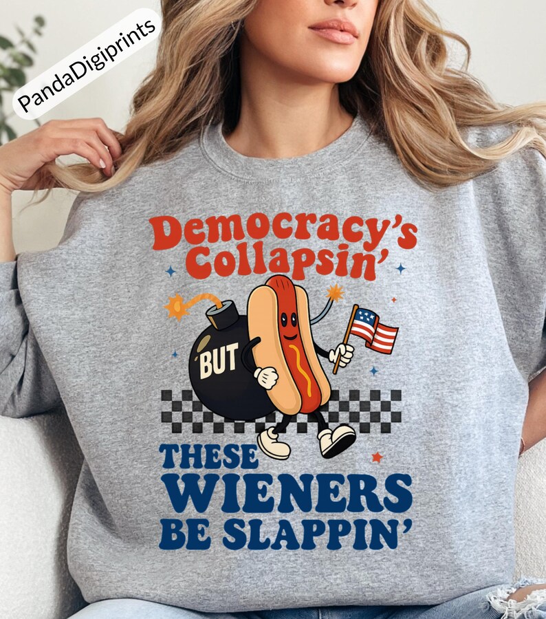 May include: A gray sweatshirt with the text "Democracy's Collapsin' BUT THESE WIENERS BE SLAPPIN'". The graphic features a cartoon hot dog holding an American flag next to a bomb.