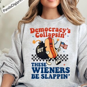 May include: A light gray sweatshirt with the text "Democracy's Collapsin' BUT These Wieners Be Slappin'". The graphic features a cartoon hot dog holding an American flag and a bomb.
