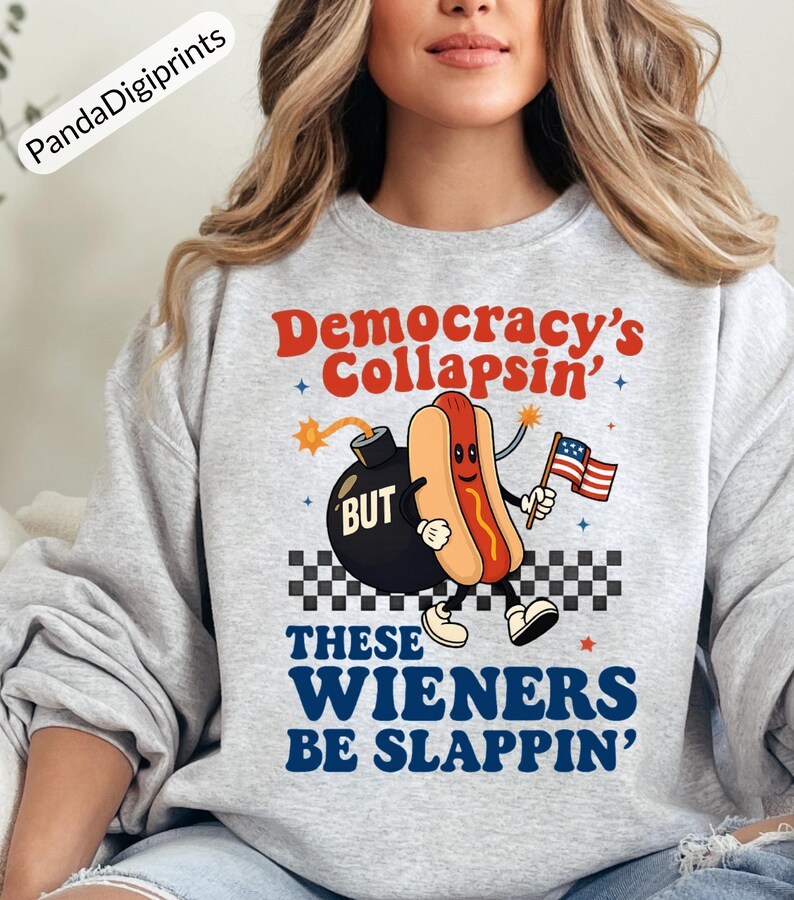 May include: A light gray sweatshirt with the text "Democracy's Collapsin' BUT These Wieners Be Slappin'". The graphic features a cartoon hot dog holding an American flag and a bomb.