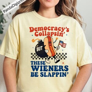 May include: A light yellow t-shirt with the text "Democracy's Collapsin' BUT These Wieners Be Slappin'". The graphic features a cartoon hot dog holding an American flag next to a bomb.