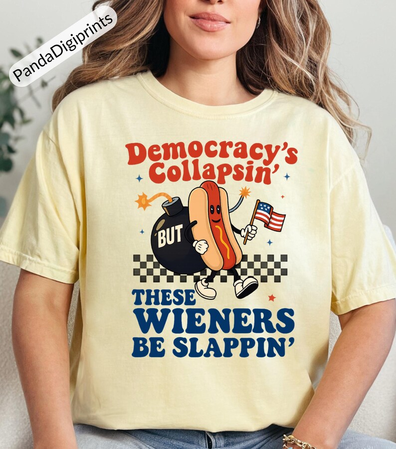 May include: A light yellow t-shirt with the text "Democracy's Collapsin' BUT These Wieners Be Slappin'". The graphic features a cartoon hot dog holding an American flag next to a bomb.