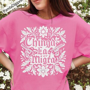 May include: A pink t-shirt featuring a white floral design and the text "Chinga La Migra". The design includes two revolvers. The shirt is a solid pink colour.