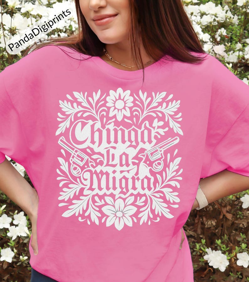 May include: A pink t-shirt featuring a white floral design and the text "Chinga La Migra". The design includes two revolvers. The shirt is a solid pink colour.