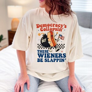 May include: Cream-colored t-shirt with the text "Democracy's Collapsin'" and "These Wieners Be Slappin'". The graphic features a cartoon hot dog holding an American flag next to a bomb.