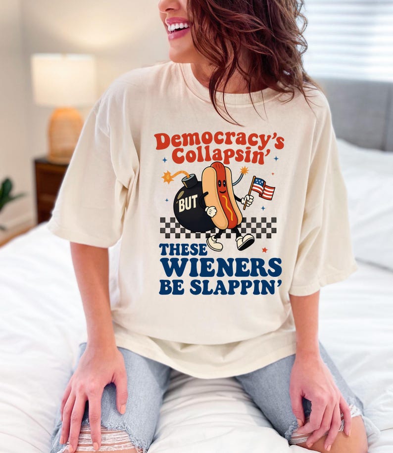 May include: Cream-colored t-shirt with the text "Democracy's Collapsin'" and "These Wieners Be Slappin'". The graphic features a cartoon hot dog holding an American flag next to a bomb.