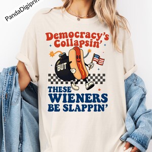 May include: Cream-colored t-shirt with the text "Democracy's Collapsin' BUT These Wieners Be Slappin'". The graphic features a cartoon hot dog holding an American flag and a bomb. The shirt has short sleeves.
