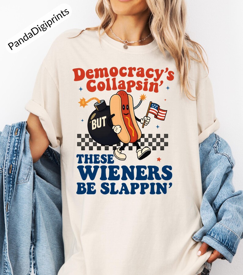 May include: Cream-colored t-shirt with the text "Democracy's Collapsin' BUT These Wieners Be Slappin'". The graphic features a cartoon hot dog holding an American flag and a bomb. The shirt has short sleeves.