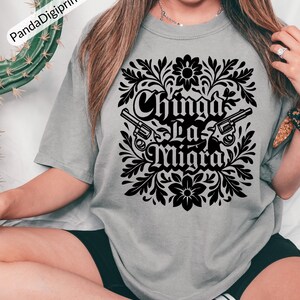 May include: A grey t-shirt with a black floral design, two revolvers, and the text "Chinga La Migra". The shirt is a relaxed, casual style.