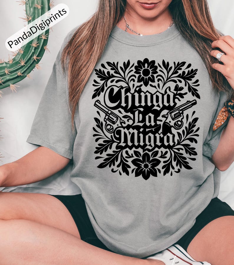 May include: A grey t-shirt with a black floral design, two revolvers, and the text "Chinga La Migra". The shirt is a relaxed, casual style.