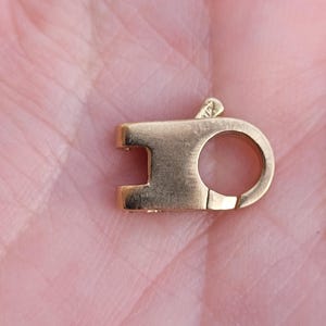 May include: A small, gold-coloured jewellery clasp with a rectangular bar and a circular loop. The clasp has a hinged design for secure closure. The metal has a polished finish, reflecting light. The clasp is likely used for necklaces or bracelets.