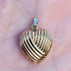 May include: A gold-colored heart-shaped pendant with a textured, ribbed design. The pendant has a small loop at the top for a chain. The heart shape is prominent, and the gold color gives it a classic look. Jewelry.