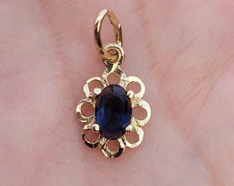 Pode incluir: Pingente dourado com uma pedra preciosa oval azul escuro no centro. A pedra preciosa é rodeada por uma moldura em forma de flor. O pingente tem uma pequena argola na parte superior para prender a uma corrente ou pulseira. Fundo neutro.