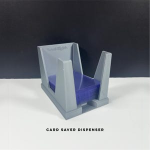 May include: A gray card saver dispenser holding a stack of blue card sleeves. The dispenser has a rectangular base with angled supports and the text "Semi-Rigid" on the side. The text "CARD SAVER DISPENSER" is printed at the bottom.