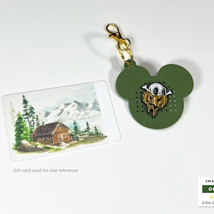 May include: An olive green mouse-shaped tag with a gold lobster clasp and a central enamel pin featuring a white skull atop a melting gold pumpkin. The tag measures 2.6 inches long by 2.5 inches wide, shown next to a gift card for scale.