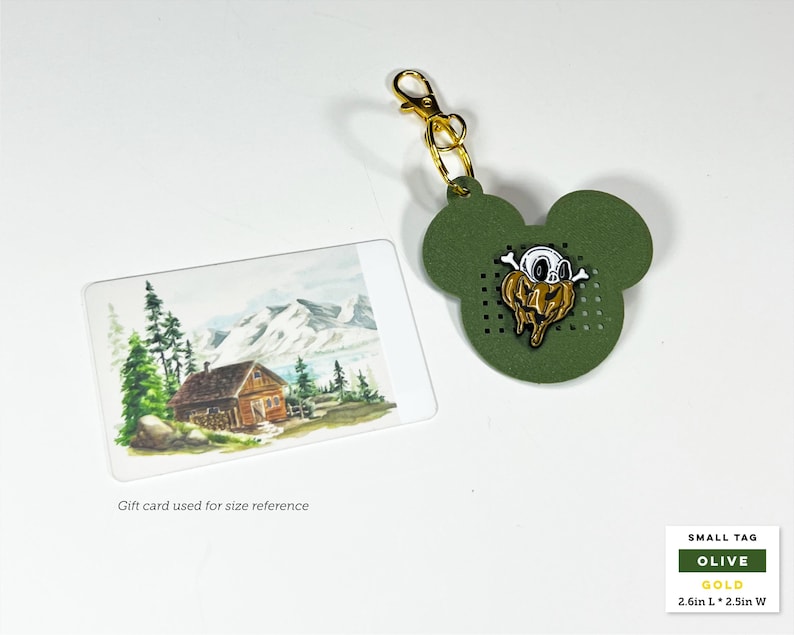 May include: An olive green mouse-shaped tag with a gold lobster clasp and a central enamel pin featuring a white skull atop a melting gold pumpkin. The tag measures 2.6 inches long by 2.5 inches wide, shown next to a gift card for scale.