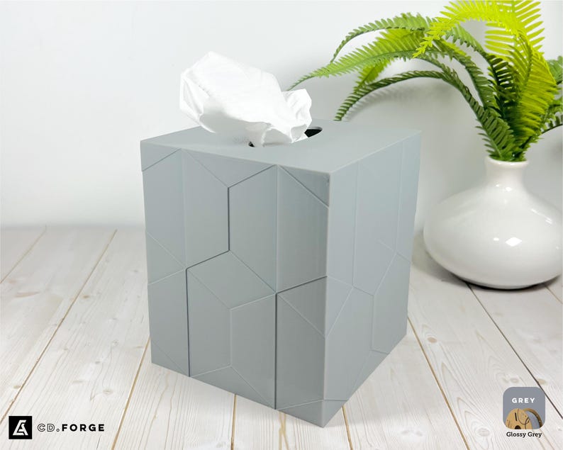 Pu&ograve; includere: Un copriscatola per fazzoletti quadrato di colore grigio chiaro con un motivo geometrico a facce sulla superficie. La scatola presenta un'apertura ovale centrale sulla parte superiore da cui sporge un fazzoletto bianco. Il testo CD.FORGE &egrave; in basso.
